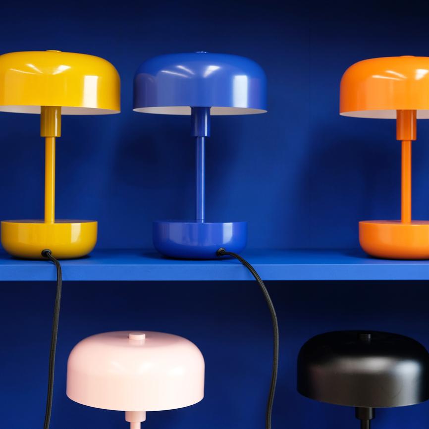 Flerfarvede bordlamper (gul, blå, orange øverst; lyserød, sort nederst) står på blå hylder; deres sorte ledninger hænger ned foran en dybblå baggrund.
