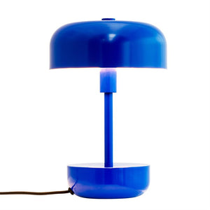 Blå, rund bordlampe lyser svagt mellem skærm og søjle; cylindrisk stang forbinder top- og bundskåle, placeret mod hvid baggrund med sort stofledning til venstre.