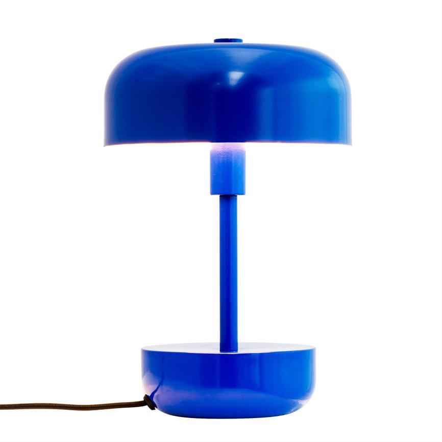 Blå, rund bordlampe lyser svagt mellem skærm og søjle; cylindrisk stang forbinder top- og bundskåle, placeret mod hvid baggrund med sort stofledning til venstre.