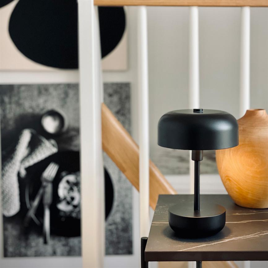 Sort bordlampe står på et marmoreret sidebord ved en trævase, foran hvidt spindelgelænder og sort-hvidt vægkunst i et lyst, moderne hjem.