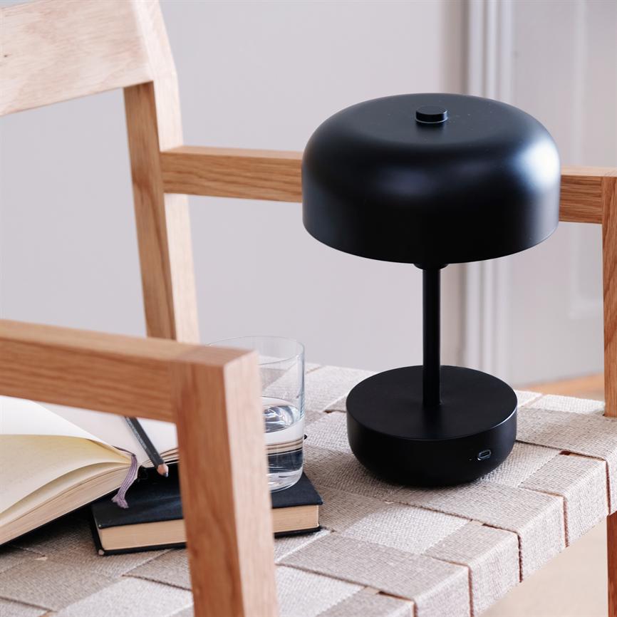 Sort, moderne bordlampe står på et flettet sæde af en træstol; ved siden af ligger notesbøger og en blyant samt et glas vand i et lyst, minimalistisk rum.