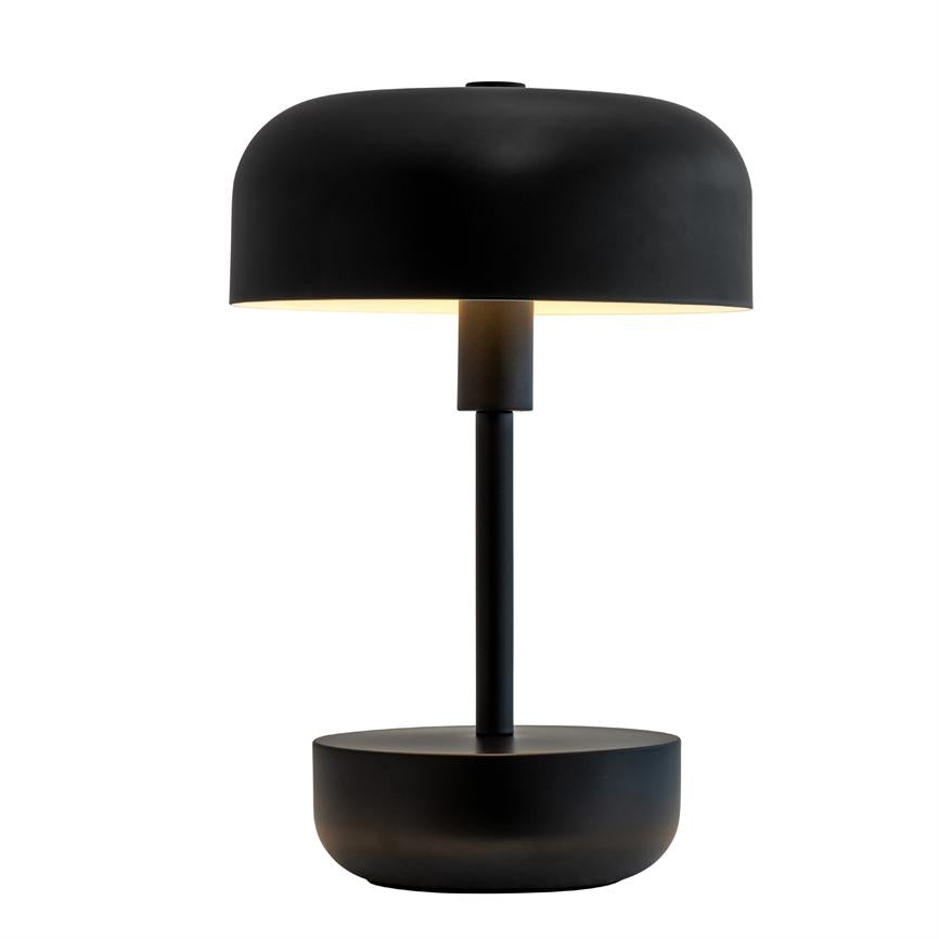 Sort bordlampe med rund, kuppelformet skærm lyser blødt nedad; den står på en slank stang og rund base mod en ren, hvid baggrund.