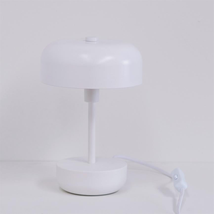 Hvid bordlampe står slukket på en hvid bordplade; rund base, tynd skaft og kupolformet skærm, med hvid ledning og vippekontakt synlig til højre.