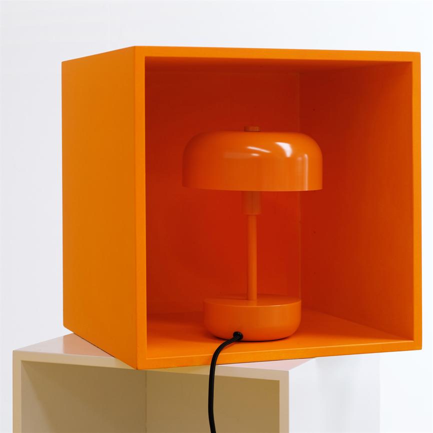 Orange bordlampe står inde i en åben orange kube, med sort ledning hængende ud foran; kuben er stablet på en hvid kube mod hvid baggrund.