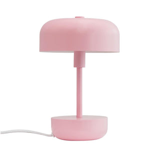 En mat lyserød bordlampe står opret; rund skærm over en smal stang og rund base, placeret mod en ensartet hvid baggrund.