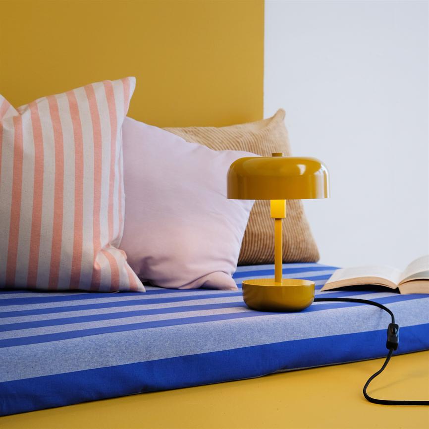 Gul bordlampe står på en blå- og lyseblå stribet hynde ved en åben bog; omkring den ligger lyserøde, stribede og beige puder foran gul og hvid baggrund.