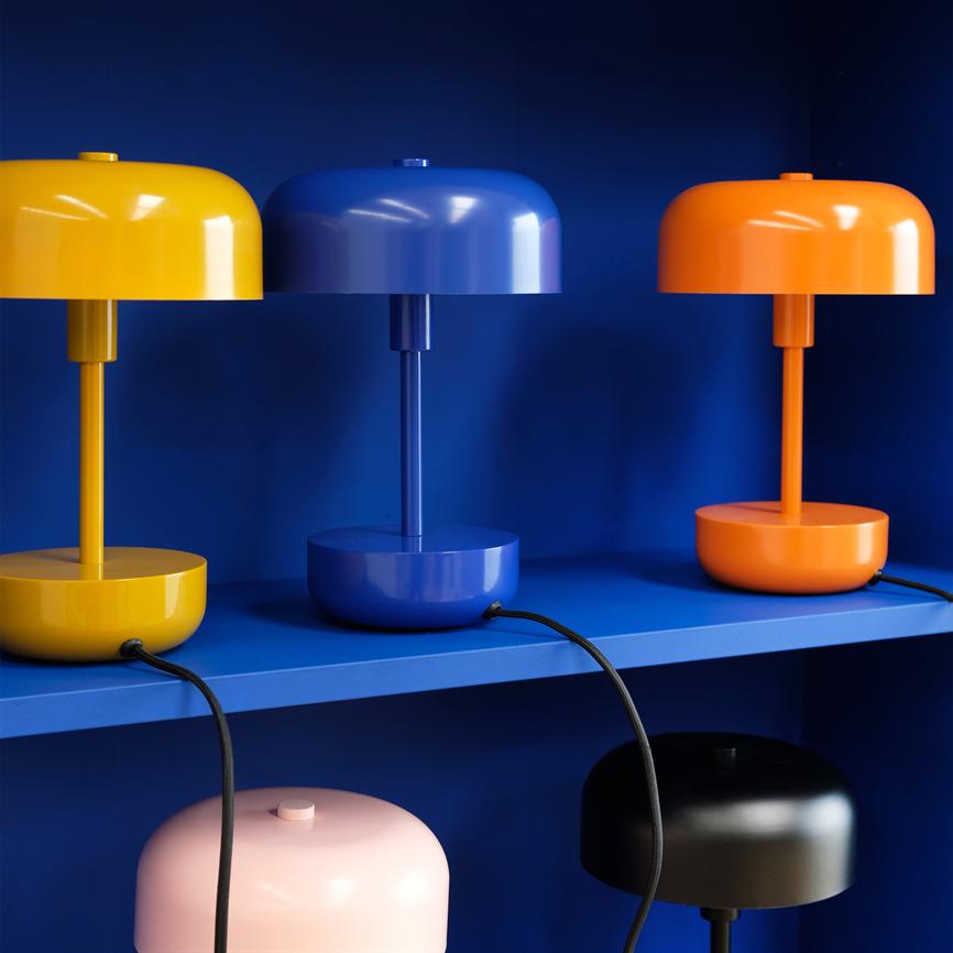 Tre glatte, runde bordlamper i gul, blå og orange står side om side på en blå hylde, tilsluttet med sorte kabler mod en dybblå baggrund.