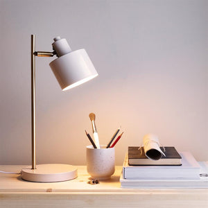 En hvid bordlampe lyser ned på en porcelænskop med blyanter og en pensel, placeret på et lyst træbord ved en stak bøger og et sammenrullet papir mod en lys væg.