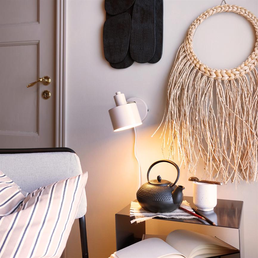 En sort støbejerns tekande står på et lille bord, oplyst af en væglampe; ved siden af en kop, åben bog og flettet vægdekoration i et hyggeligt stuehjørne.