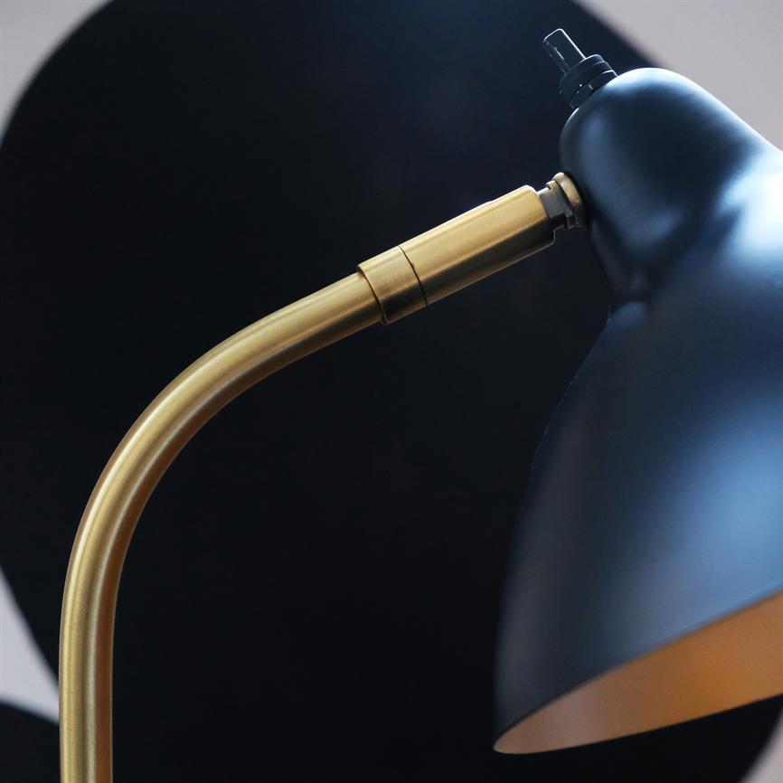 Skrivebordslampe med sort skærm og buet, messingfarvet arm peger nedad foran en mørk, cirkelformet baggrund i et minimalistisk interiør.