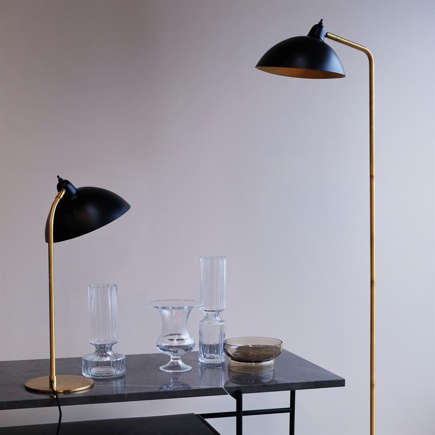En bordlampe og en gulvlampe i sort og messing bøjer deres skærme ned over et mørkt marmorbord, hvor flere klare glasvaser og en røget glasskål står foran en lys, tom væg.