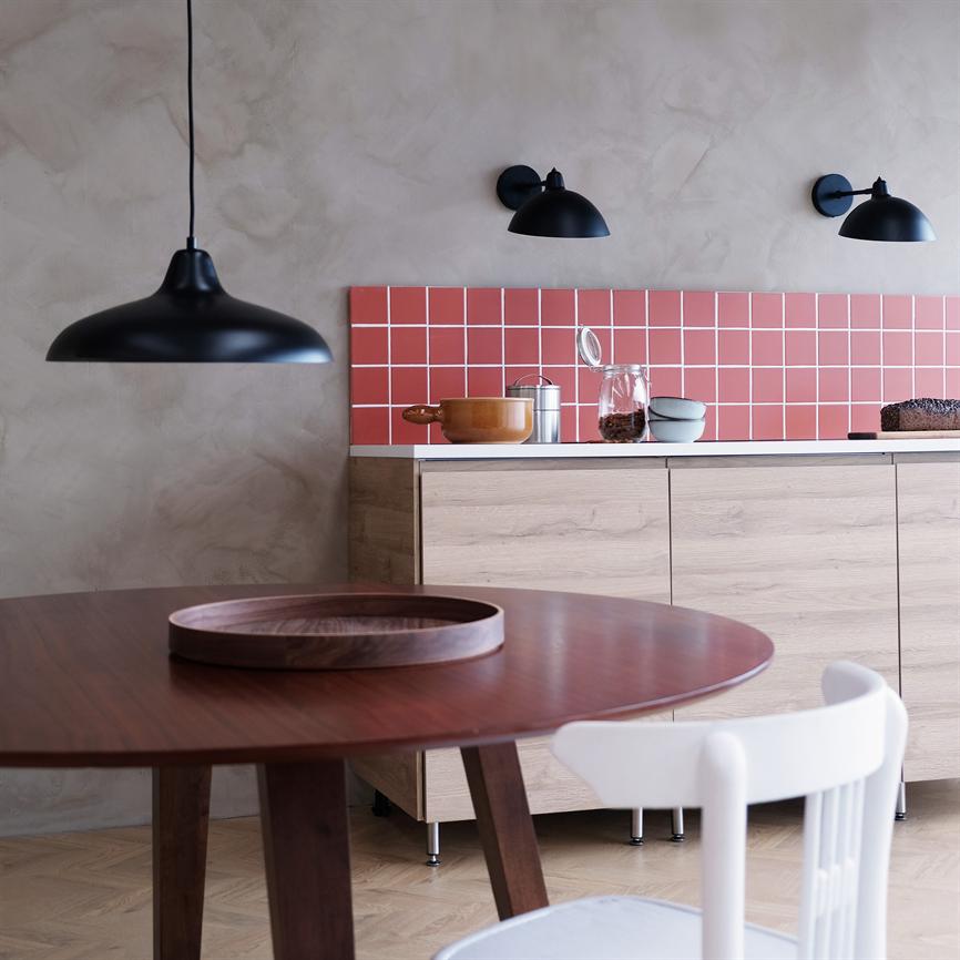 Rundt træbord med et rundt fad står i forgrunden; i baggrunden er et lyst skab med røde fliser, køkkengrej og tre sorte lamper på en sandfarvet væg.