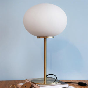 Bordlampe med stor hvid glaskuppel står på en messingstang, placeret på en bunke bøger på et træbord, foran en lys blå væg.