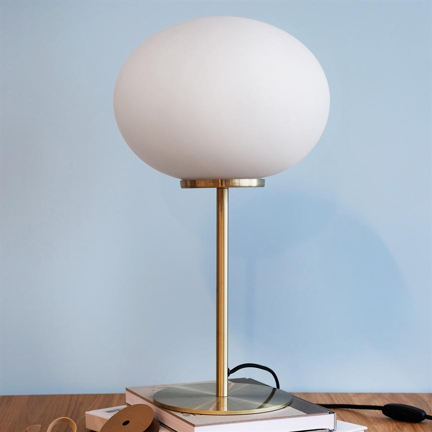 Bordlampe med stor hvid glaskuppel står på en messingstang, placeret på en bunke bøger på et træbord, foran en lys blå væg.