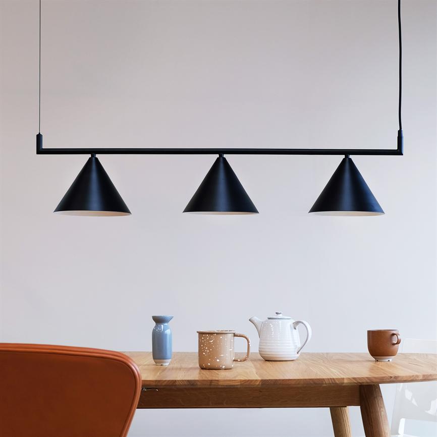Tre sorte koniske pendellamper hænger fra en vandret stang over et træbord. På bordet står en hvid tekande, tre krus og en lille blå vase; et brunt læderstolryg skimtes.