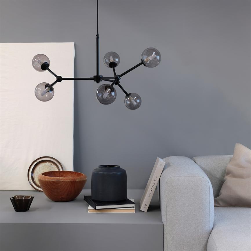 En sort, molekylær lysekrone hænger over et gråt skænk; ved siden af ligger træskåle, en sort vase og en bog med rygtekst "THE KINFOLK HOME", foran en lysegrå sofa.