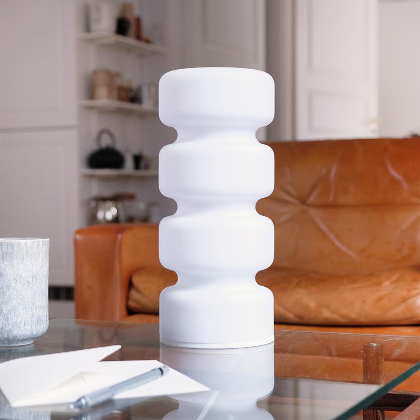 Hvid, bølget bordlampe står på et glasbord; omgivet af en notesblok og kuglepen i forgrunden, foran en brun lædersofa og hylder i en hyggelig stue.