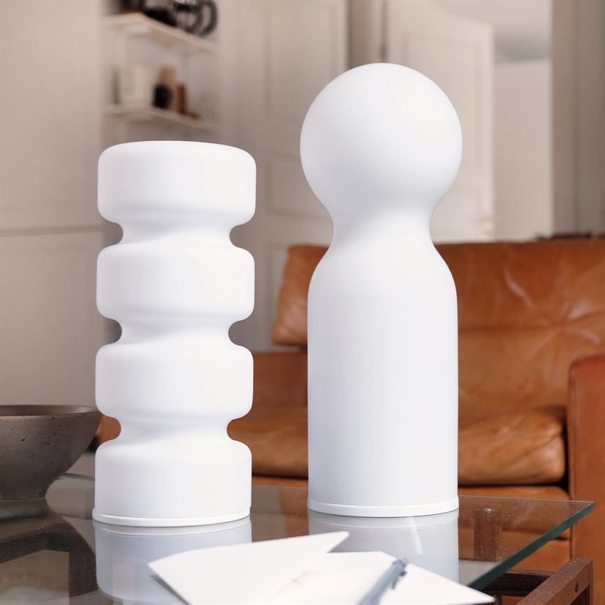 To hvide, skulpturelle vaser står side om side på et glas-sofabord; den ene er bølget, den anden rundtopet, foran en brun lædersofa i et lyst opholdsrum.