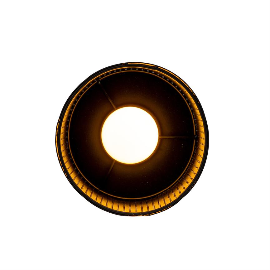 En rund loftlampe afgiver intenst varmt lys fra centrum, omgivet af en mørk, perforeret inderskærm, som kaster en gylden glød mod en hvid baggrund.