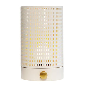 Cylindrisk bordlampe med hvid perforeret metalhylster; varmt lys skinner gennem rektangulære huller, monteret på mat hvid fod med rund, messingfarvet drejeknap, placeret mod hvid baggrund.