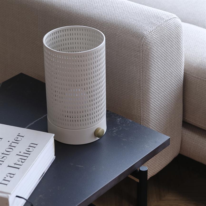 Hvid perforeret cylinderlampe står på et sort sidebord ved en beige sofa; en bog ved siden af viser teksten: "Historier Tra Italien".