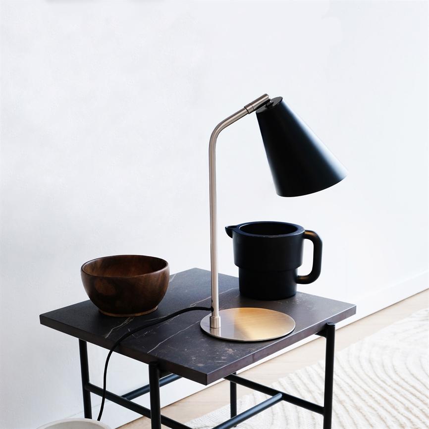 Skrivebordslampe kaster lys ned på et lille sidebord med en træskål og et sort krus; minimalistisk lyst rum med hvid væg og lyst trægulv.