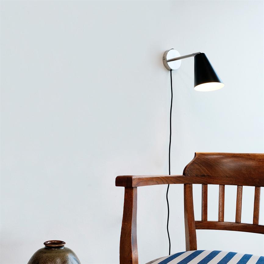Sort kegleformet væglampe lyser ned over en træstol med lodrette ryglister og blå‑hvid stribet hynde, hængende på en hvid væg med synlig sort ledning; en keramisk krukke på gulvet.