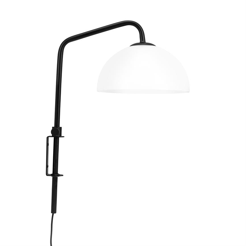 Væglampe med sort metalarm og klemme holder en halvkugleformet hvid skærm; armen bøjet udad, ledning hænger ned, mod en ensartet hvid baggrund.