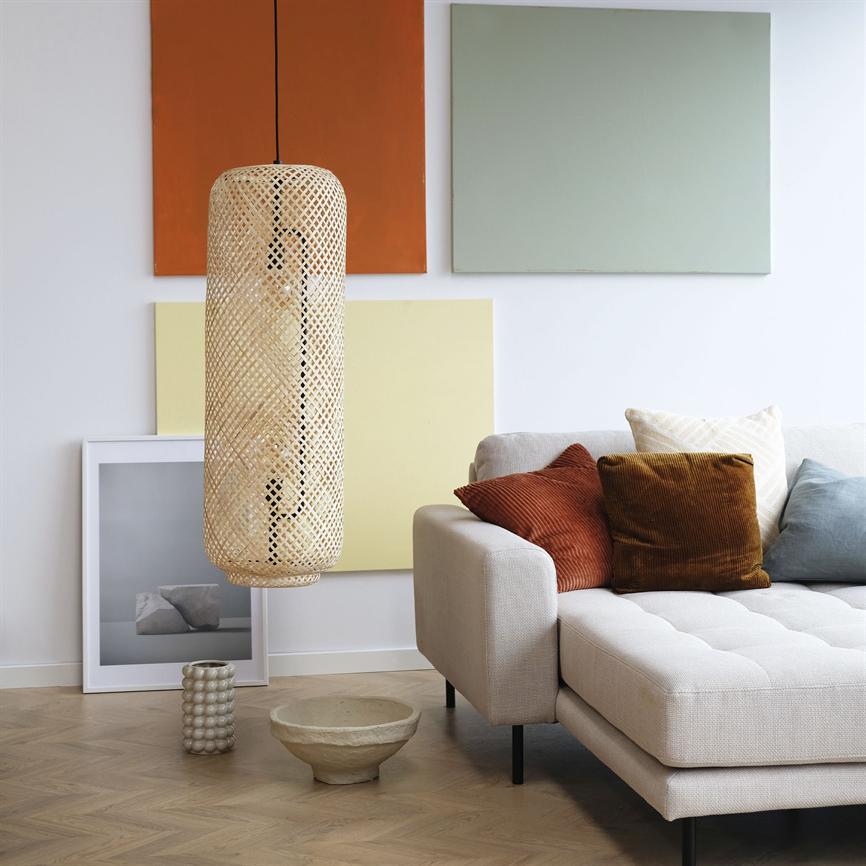 En cylindrisk flettet rattanpendel hænger foran farvede lærreder over et lyst stuegulv; ved siden af står en beige sofa med rust-, brun- og blå puder og keramiske skåle.