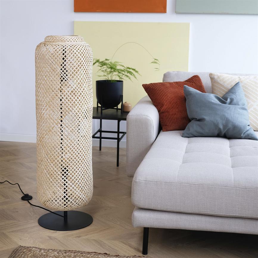 En høj flettet gulvlampe står ved siden af en lys sofa og kaster blødt lys i en moderne stue med farvede puder, sort sidebord med plante og lyst trægulv.