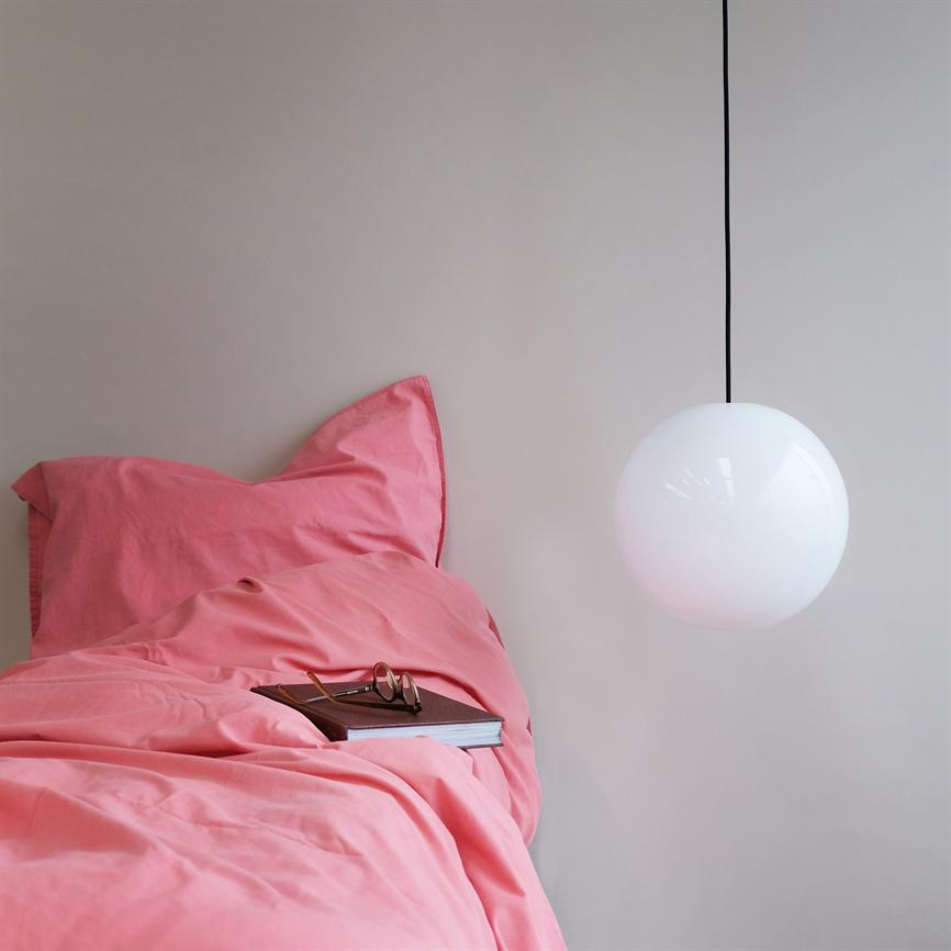 Et pink sengesæt ligger krøllet på en seng, med en bog og et par briller placeret ovenpå; en rund, hvid pendel hænger foran en neutral væg og danner enkel baggrund.