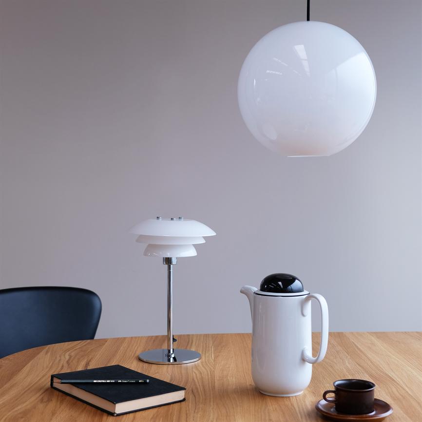 Hvid kugleformet loftslampe hænger over et lyst træbord; på bordet står en hvid termokande, en lille mørk kaffekop, en notesbog og en moderne bordlampe i et minimalistisk rum.