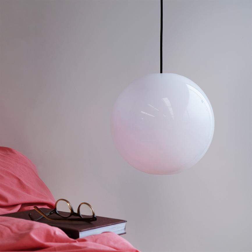 Hvid rund pendellampe hænger stille og udsender blødt, rosa-tonet lys; over en seng med pink sengetøj, hvor en lukket bog med briller ligger på sengekanten.