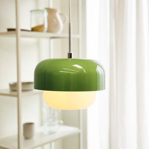 En grøn loftlampe hænger og udsender varmt lys i et lyst rum med hvide gardiner og en åben hylde med glas og keramik i baggrunden.