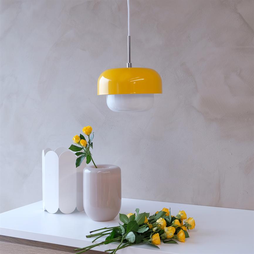 Gul pendellampe hænger over et hvidt bord; to vaser og en buket gule roser ligger på bordet i et lyst, minimalistisk rum med lyse pudsede vægge.