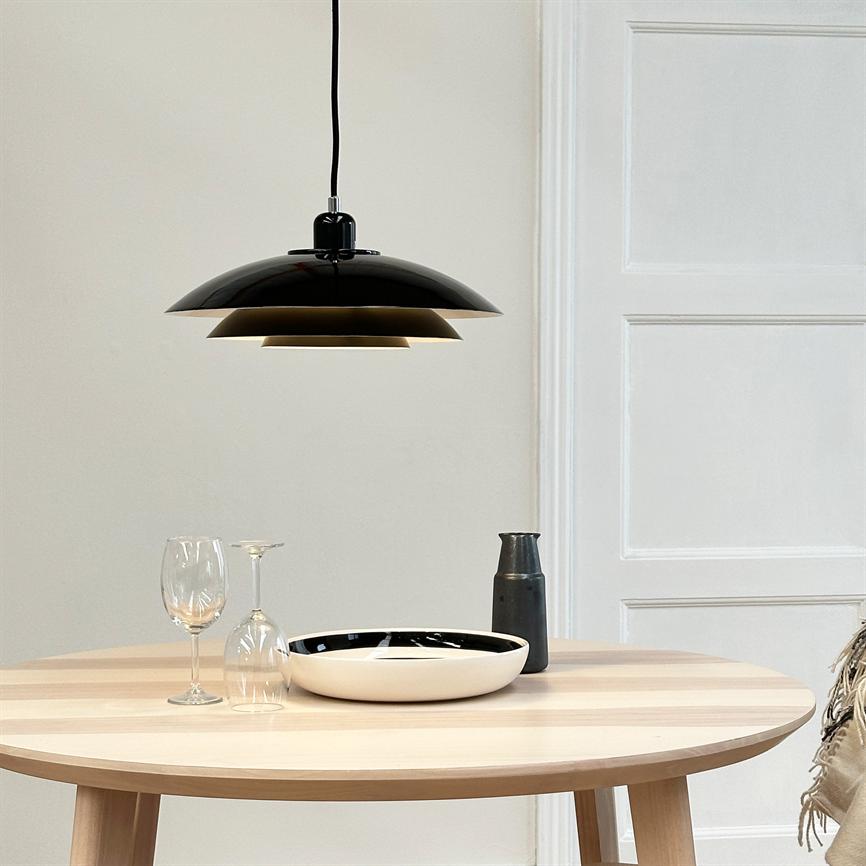 Sort pendellampe hænger over et rundt træbord; bordet har en hvid skål med sort inderside, en mørk kande og to vinglas (et oprejst, et på hovedet) i et lyst rum.