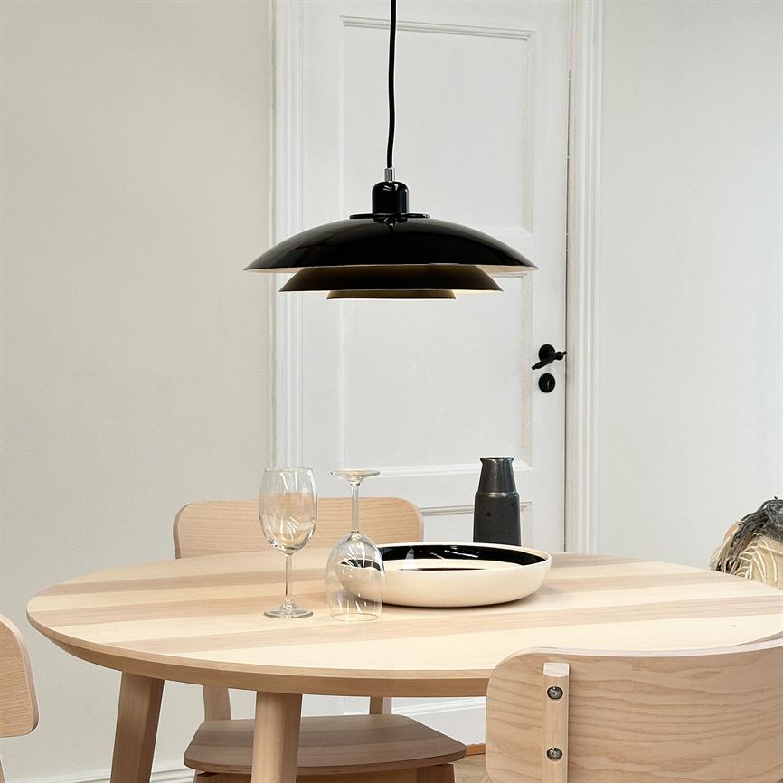 Sort pendel hænger over et rundt lyst træbord; på bordet står et opret vinglas, et vendt vinglas, en sort-hvid skål og en mørk karaffel i et lyst, minimalistisk spiserum.