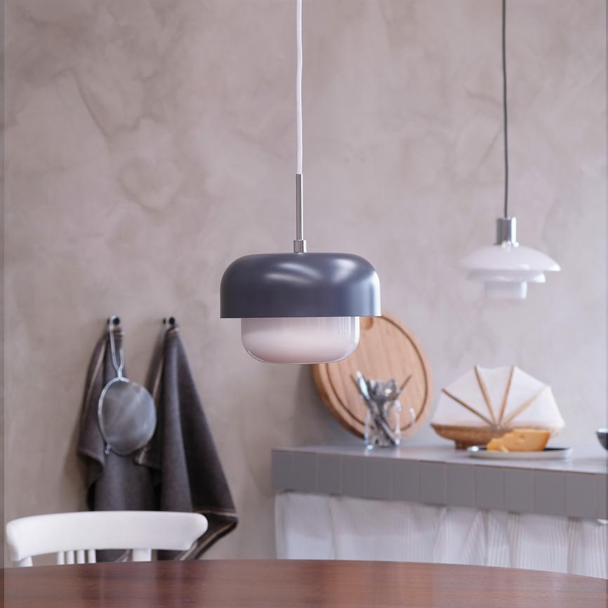 En grå pendel-lampe hænger over et rundt træbord; i baggrunden køkkenbord med skærebræt, bestik i glas, ost og servietfoldning, samt karklude hængende på væggen.