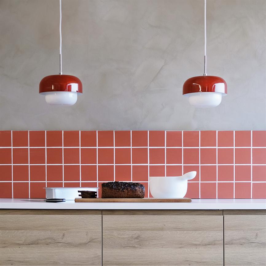 Et mørkt rugbrød ligger skåret på et skærebræt; to røde pendellamper hænger over et hvidt bord foran røde kvadratiske fliser og en grå væg, med lyse træskuffer under.