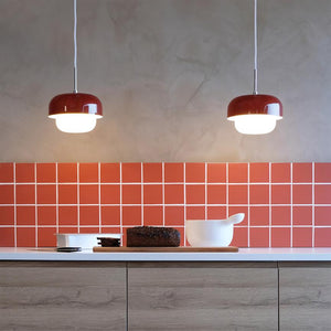 To røde pendellamper lyser ned over et køkkenbord, hvor et rugbrød ligger på et skærebræt ved siden af en hvid skål; bagvæggen er dækket af røde kvadratiske fliser.