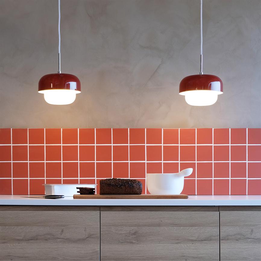 To røde pendellamper lyser ned over et køkkenbord, hvor et rugbrød ligger på et skærebræt ved siden af en hvid skål; bagvæggen er dækket af røde kvadratiske fliser.