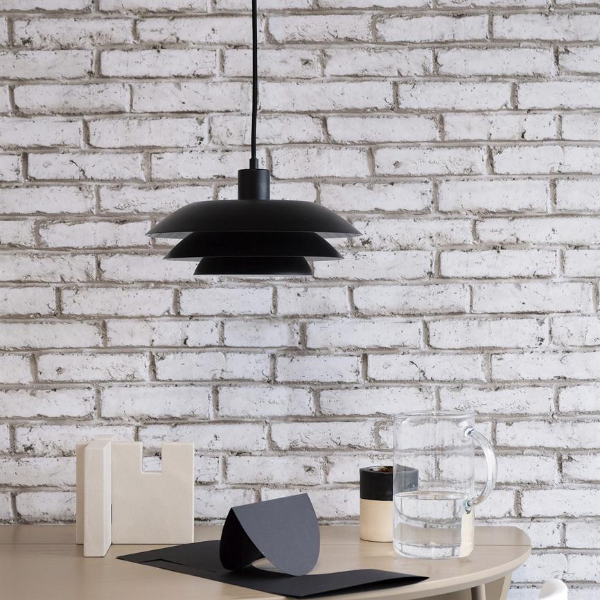 Sort lagdelt loftslampe hænger over et lyst træbord med en glaskande, en lille krukke, papirstykker og to beige dekorative blokke foran en hvid, let slidt murstensvæg.