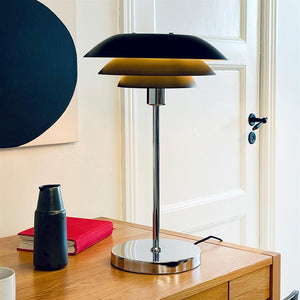 Bordlampe i krom med tre skærme lyser varmt, står på et træskrivebord ved en hvid dør, flankeret af en rød bog, en sort kande og abstrakt vægkunst.