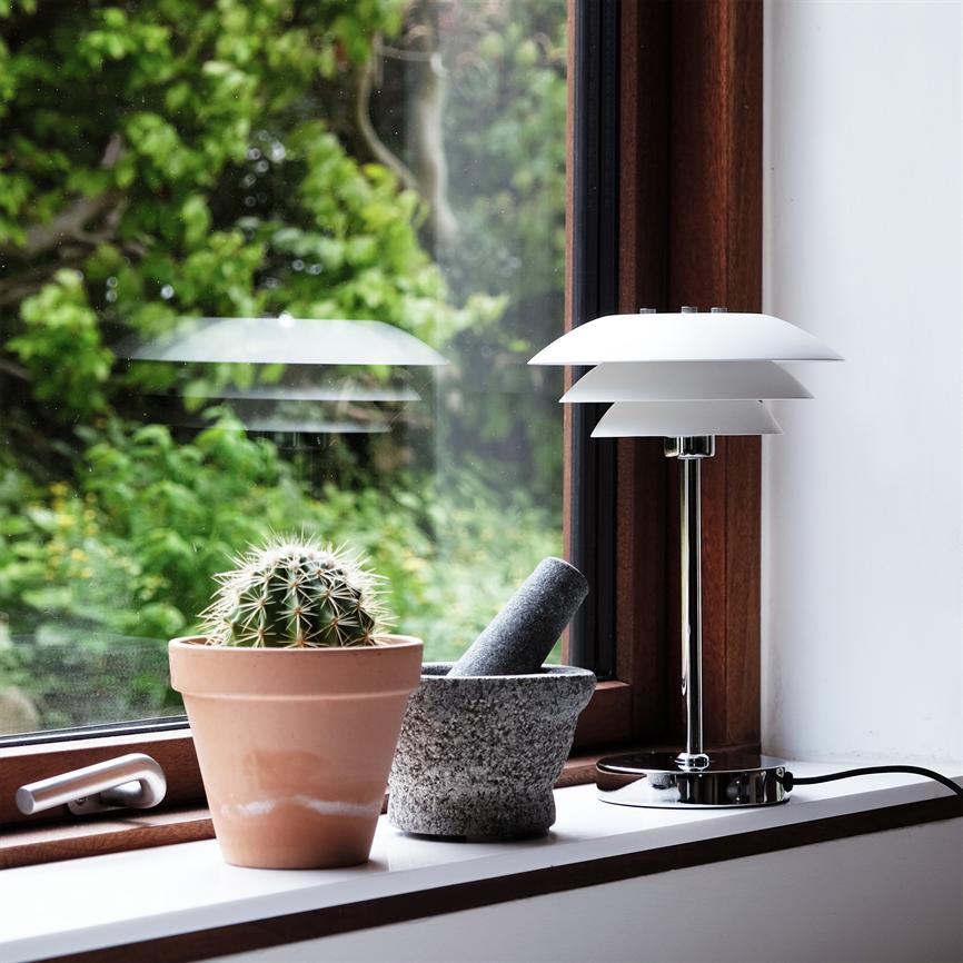 Kaktus i terrakottakrukke står på vindueskarm, flankeret af en stenmorter med støder og en hvid moderne bordlampe; grønt, sløret haveudsyn udenfor.