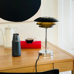 En sort-metallisk bordlampe står på et egetræsskrivebord ved en glas kop kaffe, en sort kande, en hvid kande og en rød bog foran en hvid væg med en sort cirkel.