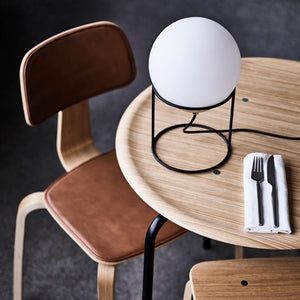 Rund bordlampe står i et sort metalstativ på et rundt lyst træbord; ledning løber henover bordet. Bestik i hvid serviet ligger ved kanten. Brun læderstol trækker sig ind.