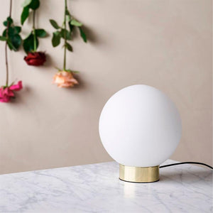 Rund hvid bordlampe står på et marmorbord med en messingfod; i baggrunden hænger tre roser — rød, fersken og pink — foran en beige væg.