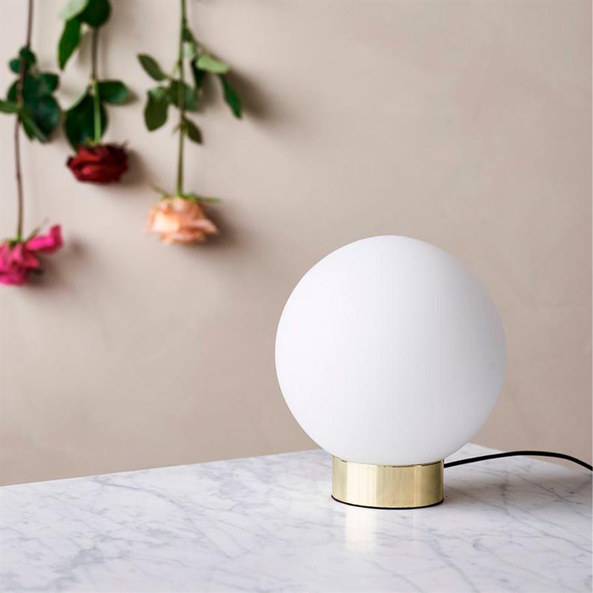 Rund hvid bordlampe står på et marmorbord med en messingfod; i baggrunden hænger tre roser — rød, fersken og pink — foran en beige væg.