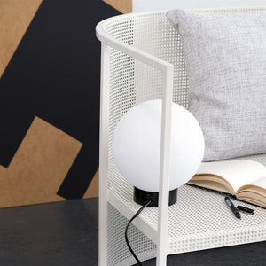 En rund hvid bordlampe står på en hvid, perforeret hylde; ved siden af ligger en åben notesbog og en pen, med en lysegrå pude og en sort‑brun plakat i baggrunden.