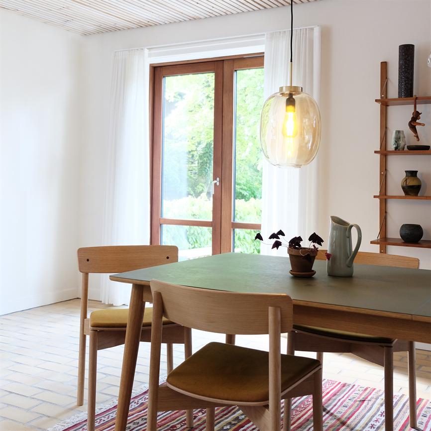 Spisebordet med fire træstole står under en stor hængende glaslampe; en kande og en lille potteplante står på bordet; store vinduer åbner til grønt haveudsnit i et lyst rum.